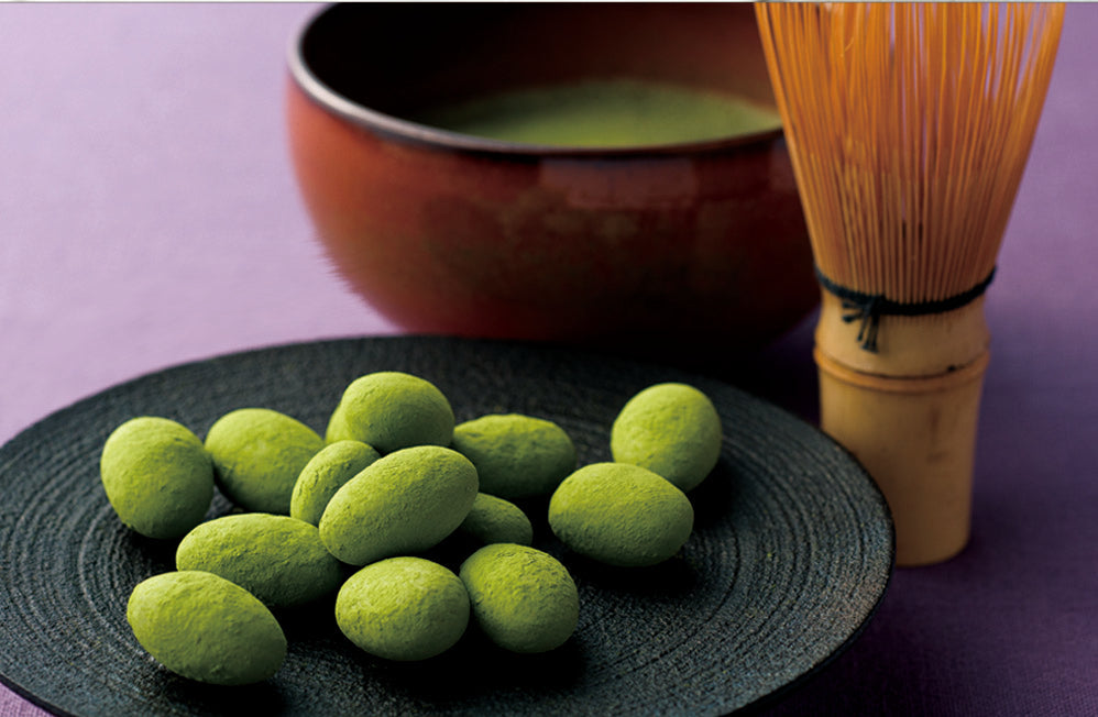 Matcha-covered chocolate almonds on plate, with traditional Japanese matcha tea whisk and bowl in background.